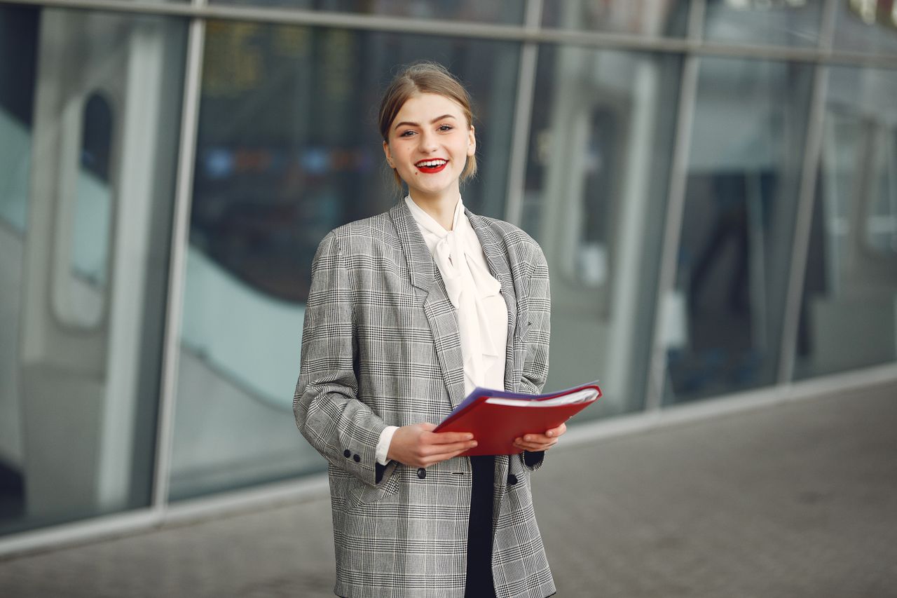 Positive young woman in trendy wear smiling at camera while holding documents standing against glass building in downtown