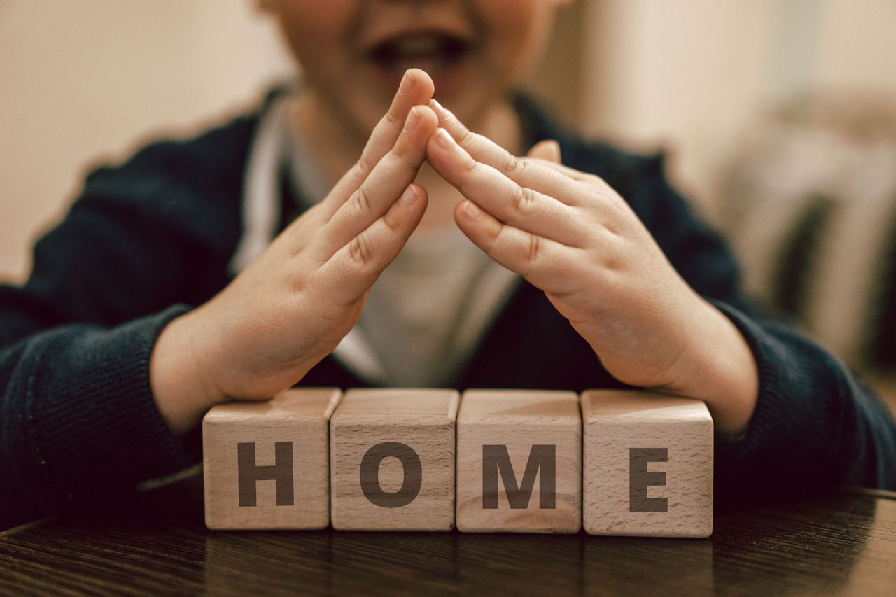 Crop excited kid with mouth opened making roof with hands above wooden cubes with word Home waiting for foster family